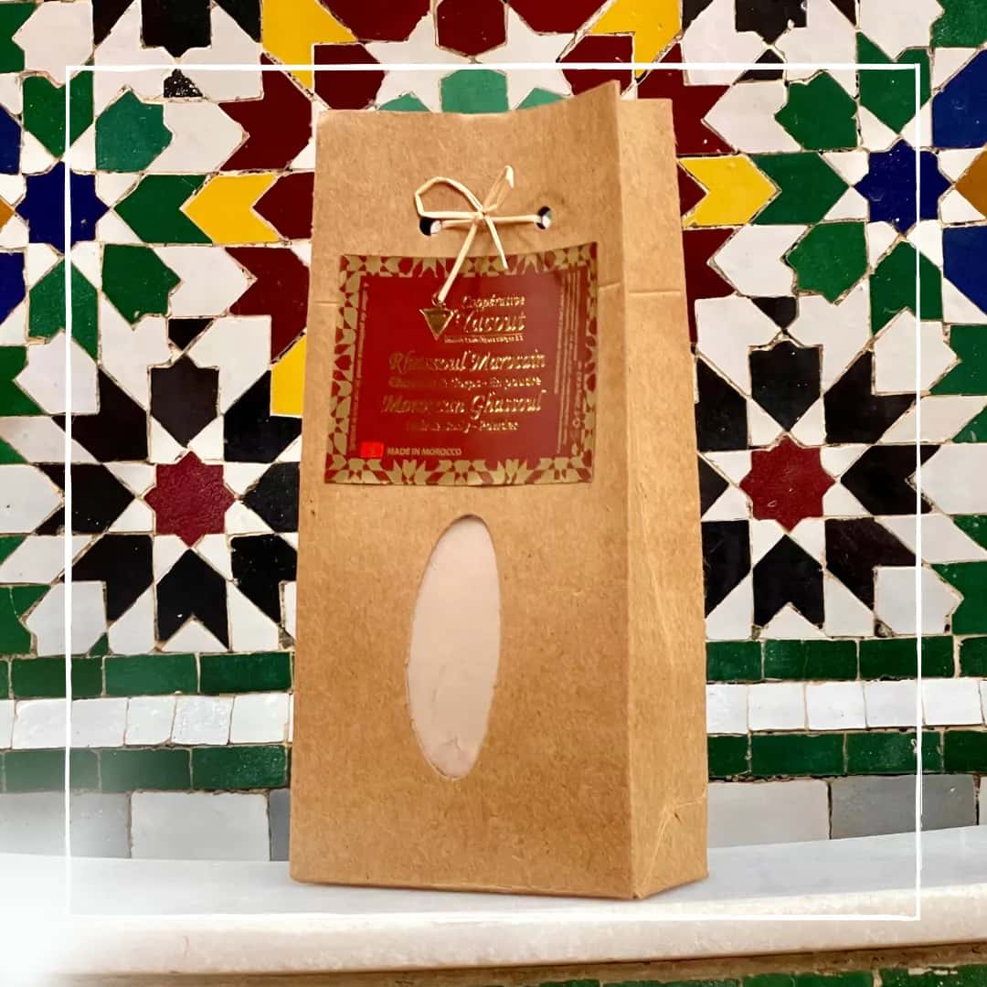 Alt text: Ghassoul marroquí puro en polvo 250g – Cooperativa Yacout, packaging artesanal kraft con ventana oval y lazo dorado, fondo de zellige tradicional marroquí