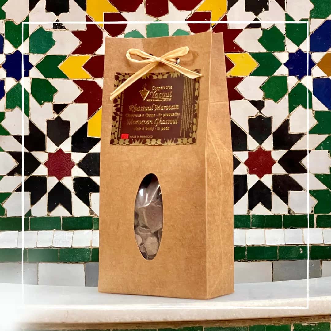 Arcilla Ghassoul Marroquí con 7 Plantas del Atlas en piedra 250g – Cooperativa Yacout, packaging kraft artesanal con lazo dorado y ventana oval, fondo de zellige tradicional marroquí`