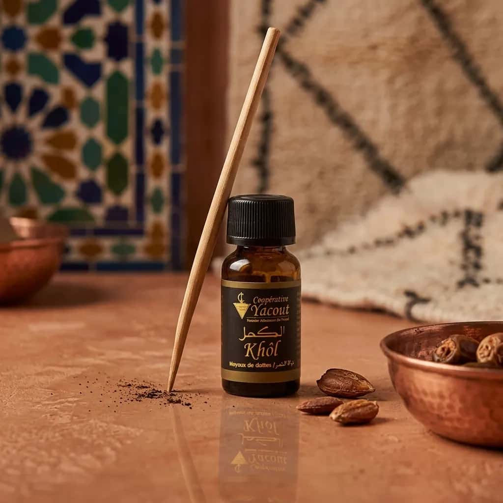 Frasco de Khôl Gris Marroquí de la marca Coopérative Yacout junto a un aplicador de madera tradicional sobre una superficie de piedra clara. La escena incluye granos sueltos de polvo de khôl, huesos de dátil y un cuenco de cobre, con un fondo decorado con azulejos marroquíes y una alfombra artesanal.