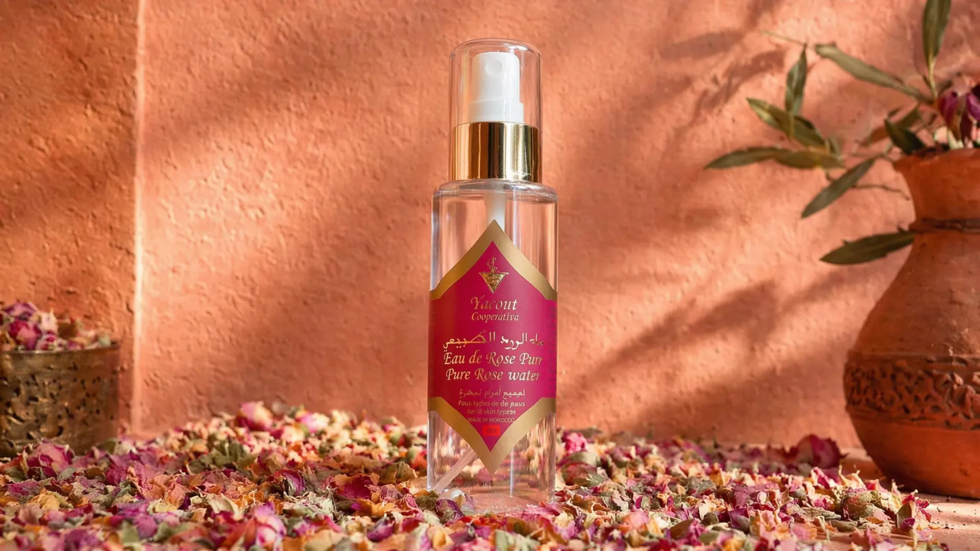 Agua de rosas pura Cooperativa Yacout 120ml sobre pétalos de rosa secos con pared terracota marroquí de fondo